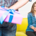 Une jeune femme souriante assise sur un canapé jaune regarde avec impatience un homme au premier plan, dont les mains floues tiennent un cadeau rose avec un nœud bleu. C'est l'occasion idéale pour surprendre une femme à coup sûr avec le bon cadeau.
