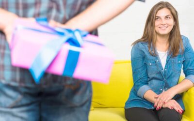 Une jeune femme souriante assise sur un canapé jaune regarde avec impatience un homme au premier plan, dont les mains floues tiennent un cadeau rose avec un nœud bleu. C'est l'occasion idéale pour surprendre une femme à coup sûr avec le bon cadeau.